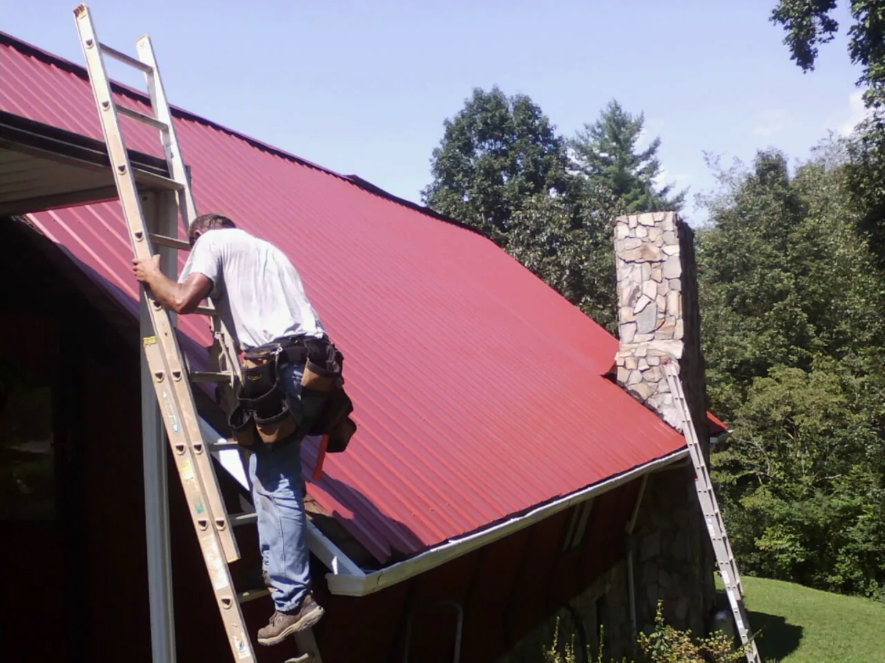 Roofer on a ladder beside a red metal roof and stone chimney — Soffit & Fascia Repair by Roofing Brothers in Saint Albans